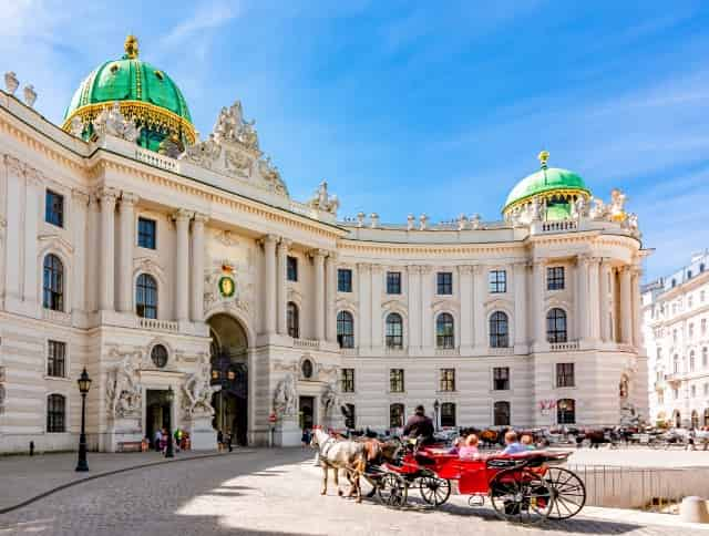 Eine rote Pferdekutsche mit Fahrgästen steht vor der Hofburg in Wien – jetzt Citytrip buchen und mit unseren Aktionscodes sparen!