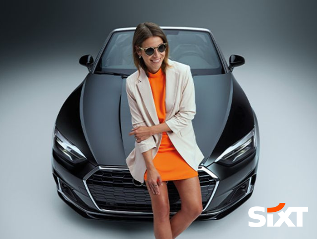 Frau in orangenem Kleid lehnt an einem Cabrio. Jetzt Mietwagen finden.