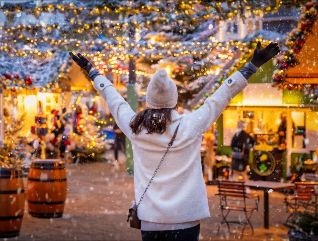 Frau steht auf einem Weihnachtsmarkt und streckt die Arme in die Luft- Jetzt deinen Flug zum Weihnachtsmarkt buchen.